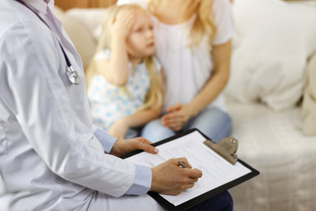 Doctor and patient. Pediatrician using clipboard while examining little girl with her mother at home. Sick and unhappy child at medical examの写真素材