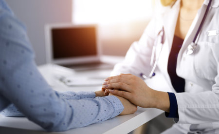 Unknown woman-doctor is holding her patient's hands to reassure the patient, discussing current health examination, while they are sitting together at the desk in the sunny cabinet in a clinic. Female physician with a stethscope at work, close-up.の写真素材