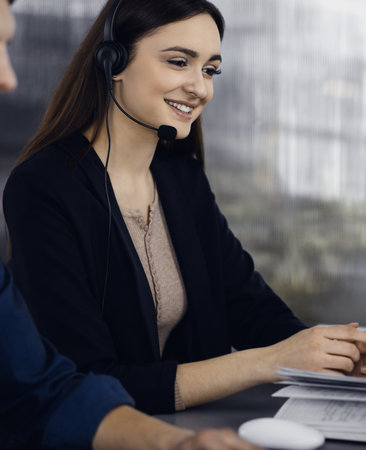 Young beautiful woman casual dressed in headsets is listening to a company's client, while she is sitting at the desk, working together with a male colleague in a modern office. Focus on woman. Call center operators at work.の写真素材