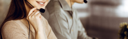 Two young people in headsets are talking to the clients, while sitting at the desk in an office. Focus on woman. Call center operators at workの写真素材