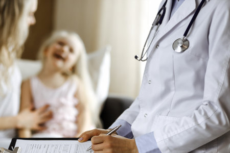 Doctor and patient. Pediatrician using clipboard while examining little girl with her mother at home. Happy cute caucasian child at medical exam. Medicine conceptの写真素材