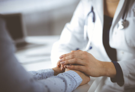 Unknown woman-doctor is holding her patient's hands, discussing current health examination, while they are sitting at the desk in the cabinet in a clinic. Physician with a stethscope at work, close-up. Perfect medical service in a hospital. Medicine concept.の写真素材