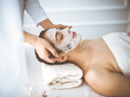 Beautiful brunette woman enjoying applying cosmetic mask with closed eyes comfortable and blissful. Relaxing treatment in medicine and spa center conceptsの写真素材