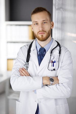 Red-bearded doctor standing straight in clinic near his working place. Portrait of physician. Medicine, healthcare