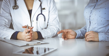 Unknown woman- doctor is listening to her patient, while sitting together at the desk in the cabinet in a clinic. Female physician with a stethoscope is writing at clipboard, close up. Perfect medical service in a hospital. Medicine concept.の写真素材