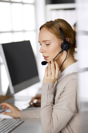 Friendly businesswoman talking by headset while sitting in office. Call center and diverse people group in businessの写真素材