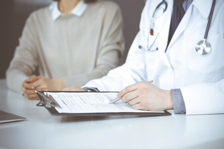 Unknown male doctor and patient woman discussing something while sitting in clinic and using clipboard. Best medical service in hospital, medicine, pandemic stopの写真素材