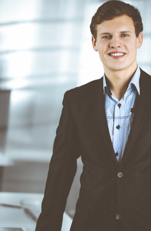 Friendly successful businessman in a black suit is standing at his workplace. Portrait of a young specialist in a modern office. Business conceptの写真素材