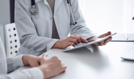 Doctor and patient sitting at the desk in clinic office. The focus is on female physicians hands using tablet computer, close up. Perfect medical service and medicine conceptの写真素材