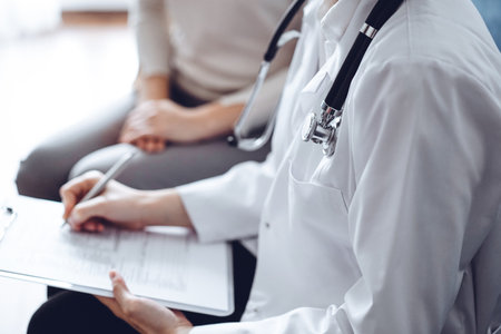 Doctor and patient sitting at sofa in clinic office. The focus is on female physicians hands filling up the medication history record form, close up. Medicine conceptの写真素材