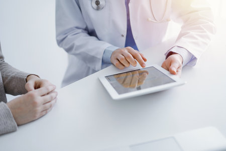 Doctor and patient sitting at the table in clinic. The focus is on female physicians hands using tablet computer, close up. Medicine and healthcare conceptの写真素材