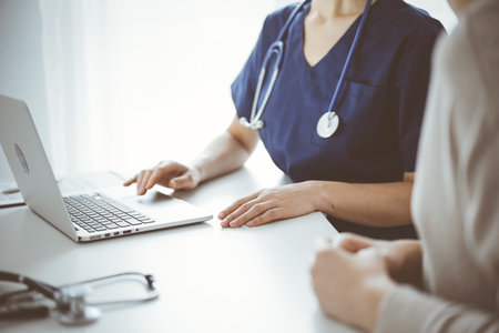Doctor and patient sitting at the desk in clinic office. The focus is on unknown female physicians hands using a laptop computer, close up. Medicine conceptの写真素材