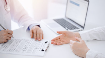 Doctor and patient discussing current health examination while sitting opposite each other at the desk in clinic. The focus is on female hands, close up. Medicine conceptの写真素材