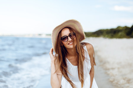 Happy smiling woman in free happiness bliss on ocean beach standing and posing with hat and sunglasses. Portrait of a female model in white summer dress enjoying nature during travel holidays vacation outdoorsの写真素材