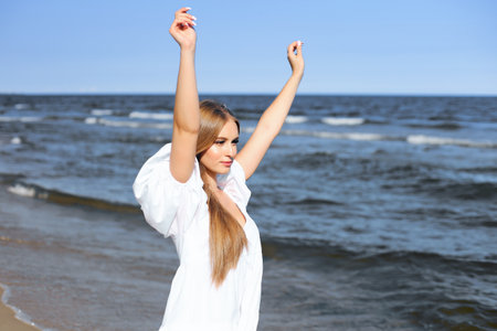 Happy, beautiful woman on the ocean beach standing in a white summer dress, raising handsの写真素材