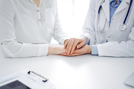 Doctor and patient sitting near each other at the table in clinic office. The focus is on female physicians hands reassuring woman, only hands, close up. Medicine conceptの写真素材