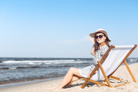Happy brunette woman wearing sunglasses and hat relaxing on a wooden deck chair at the ocean beachの写真素材