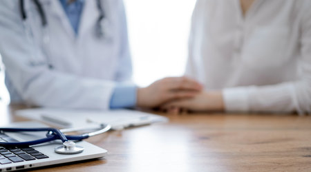 Stethoscope and laptop computer are lying on the wooden table while doctors hands reassuring a patient at the background. Medicine conceptの写真素材