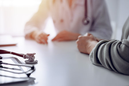 Doctor and patient are sitting and discussing something at the desk in the clinic office. The focus is on the stethoscope lying on the table, close up. Perfect medical service and medicine conceptの写真素材