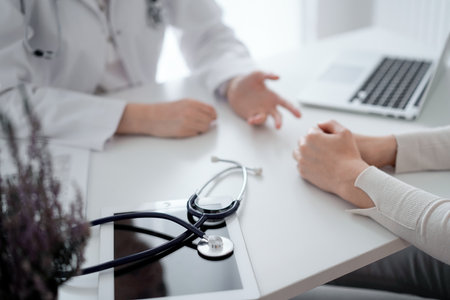 Doctor and patient sitting and discussing something at the desk in clinic. The focus is on the stethoscope lying on the table, close up. Medicine and healthcare conceptの写真素材