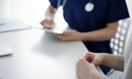 Doctor and patient sitting at the table in clinic while discussing something and using tablet computer. The focus is on female patients hands, close up. Medicine conceptの写真素材