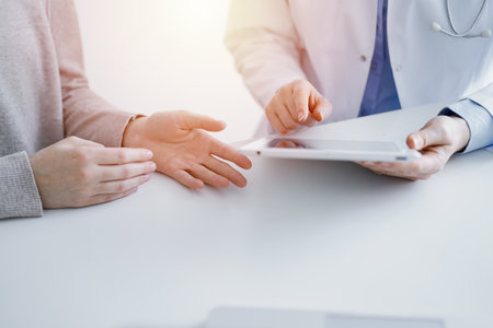Doctor and patient sitting at the table in clinic. The focus is on female physicians hands using tablet computer, close up. Medicine and healthcare conceptの写真素材