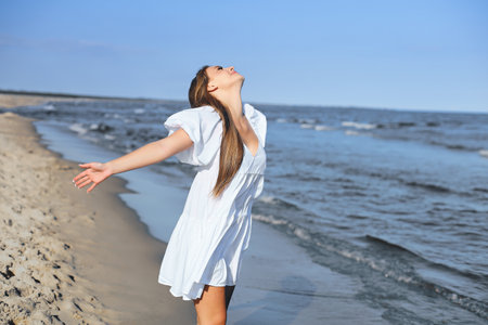Happy smiling beautiful woman is on the ocean beach in a white summer dress, open armsの写真素材