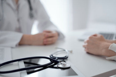 Doctor and patient sitting and discussing something at the desk in clinic. The focus is on the stethoscope lying on the table, close up. Medicine and healthcare conceptの写真素材