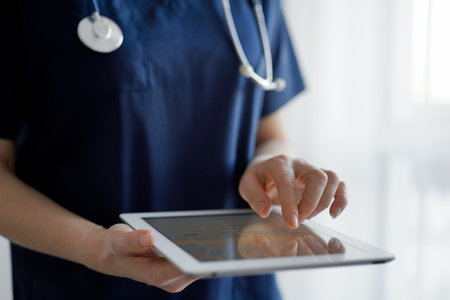 Doctor woman using tablet computer while standing near panorama window in clinic, close up. Physician or surgeon at work. Medicine conceptの写真素材