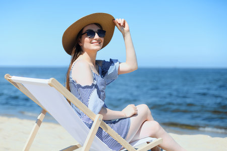 Happy young brunette woman relaxing on a wooden deck chair at the ocean beach while smiling, and wearing fashion hat, sunglasses. The enjoying vacation conceptの写真素材