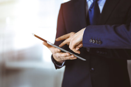 Business people use a tablet computer for discussion of their new project, standing in sunny modern office. Unknown businessman or male entrepreneur with a colleague at workplaceの写真素材