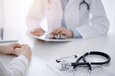 Stethoscope lying on the tablet computer in front of a doctor and patient sitting opposite each...