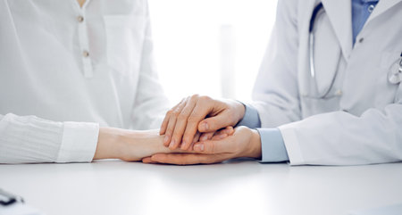 Doctor and patient sitting near each other at the table in clinic office. The focus is on female physicians hands reassuring woman, only hands, close up. Medicine conceptの写真素材