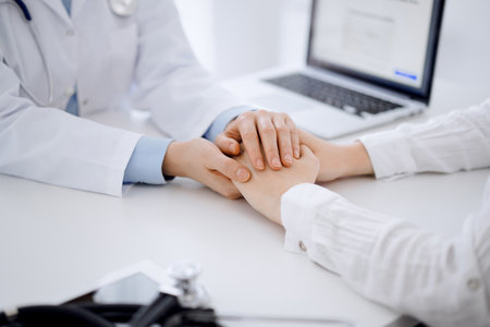 Doctor and patient sitting opposite each other at the table in clinic office. The focus is on female physicians hands reassuring woman, only hands, close up. Medicine conceptの写真素材