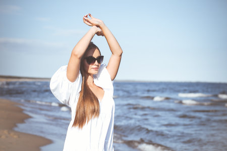 Happy blonde woman is on the ocean beach in a white dress and sunglasses, raising handsの写真素材