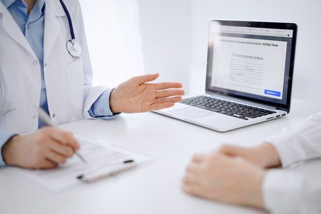 Doctor and patient sitting opposite each other at the desk in clinic. The focus is on female physicians hands pointing into laptop computer monitor, close up. Medicine conceptの写真素材
