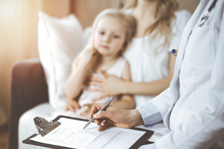 Doctor and patient. Pediatrician using clipboard while examining little girl with her mother at home. Sick and unhappy child at medical examの写真素材