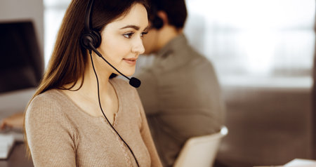 Young friendly girl in headsets is talking to a firms client, while sitting at the desk in a modern office together with her colleague. Call center operators at workの写真素材