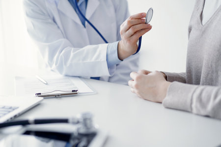 Doctor and patient sitting at the table in clinic. The focus is on female physicians hands examining woman with stethoscope, close up. Medicine conceptの写真素材