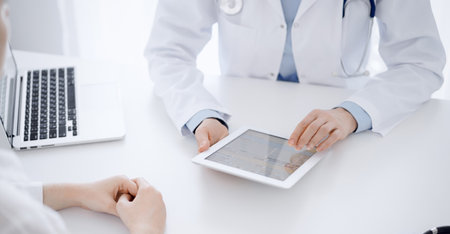 Doctor and patient sitting opposite each other at the desk in clinic. The focus is on female physicians hands pointing into tablet computer touchpad, close up. Medicine conceptの写真素材