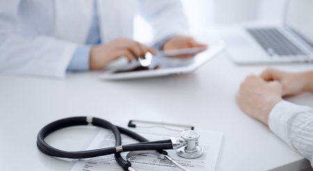 Stethoscope lying on the tablet computer in front of a doctor and patient sitting opposite each...