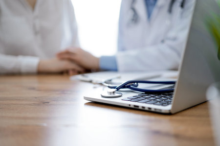 Stethoscope and laptop computer are lying on the wooden table while doctors hands reassuring a...
