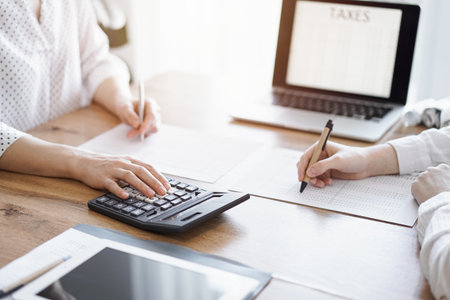 Two accountants using a laptop computer and calculator while counting taxes at wooden desk in office. Teamwork in business audit and financeの写真素材