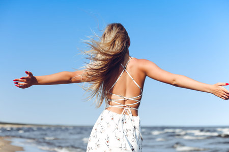 Happy blonde beautiful woman on the ocean beach standing in a white summer dress, raising hands