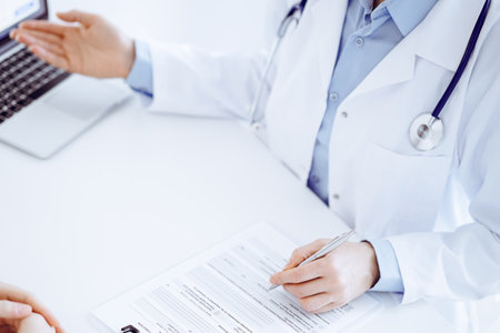 Doctor and patient sitting opposite each other at the desk in clinic. The focus is on female physicians hands pointing into laptop computer monitor, close up. Medicine conceptの写真素材