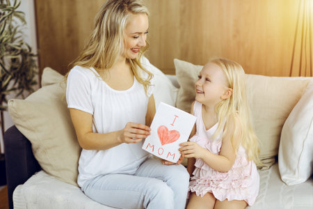 Happy mother day in sunny flat. Child daughter congratulates mom and gives her postcard with heart drawing. Family conceptの写真素材