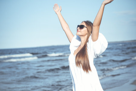 Happy blonde woman is on the ocean beach in a white dress and sunglasses, raising handsの写真素材