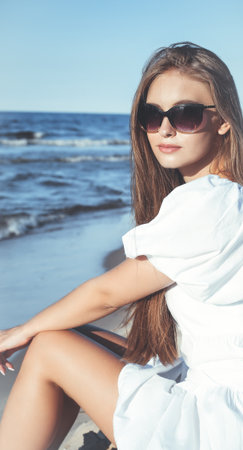 Happy, beautiful blonde woman is sitting on the ocean beach in a white summer dress and sunglassesの写真素材