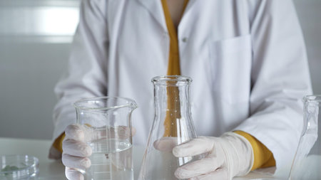A scientist with a lab coat and white protective gloves is carefully pouring a liquid from one...