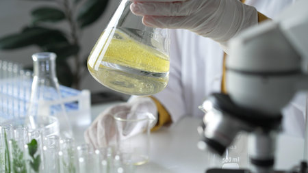 Unknown scientist wearing white protective gloves is carefully shaking up a yellow chemical solution inside an Erlenmeyer flask in a laboratory, close up. Science and medicineの写真素材